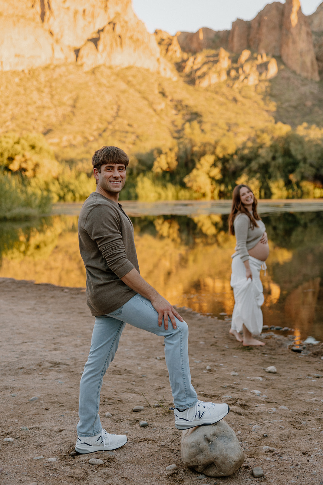 Playful maternity moment with couple by the river and desert mountains behind them in Arizona
