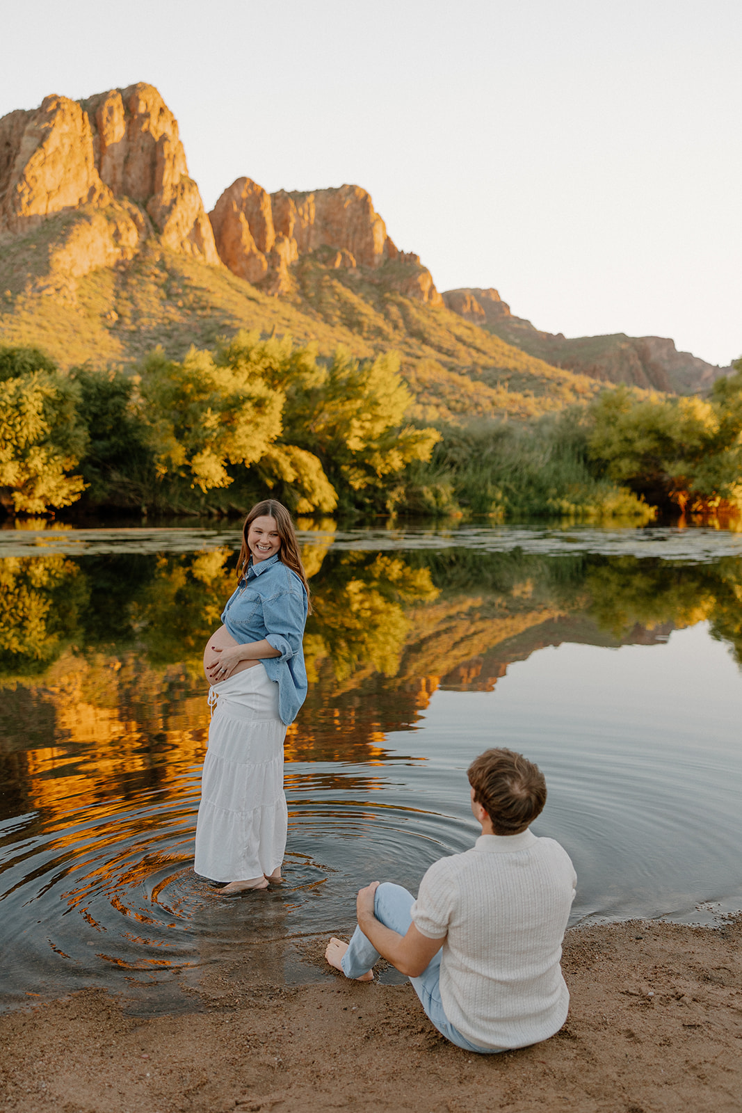 Pregnant woman standing in shallow river water while partner sits nearby, relaxed maternity session in Arizona
