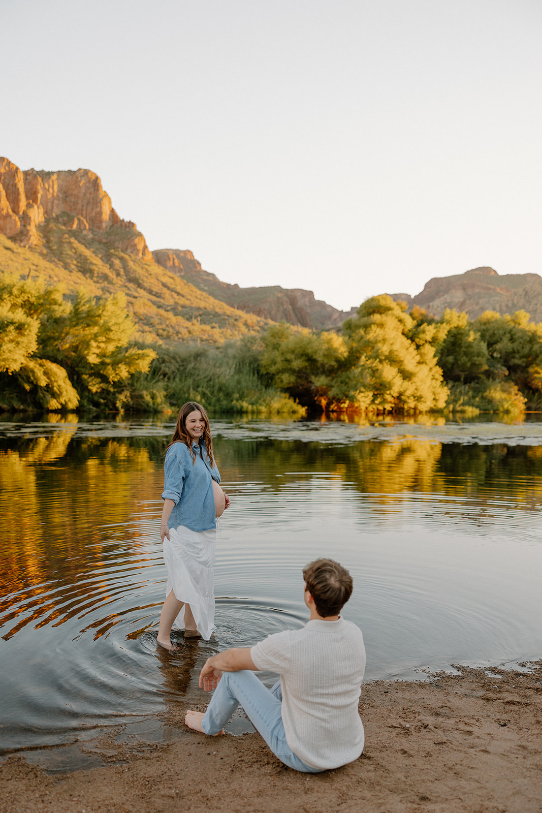 Pregnant woman standing in the river while partner sits nearby during a relaxed maternity session, Arizona maternity photographer
