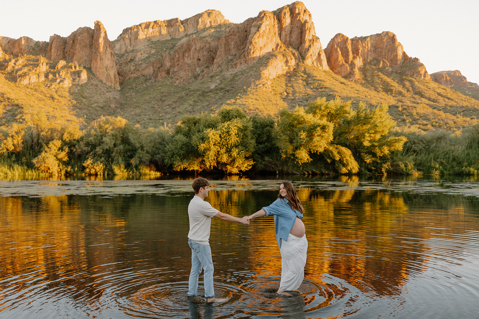 Wide shot of couple holding hands in the river with mountain reflections at sunset, Arizona maternity photographer
