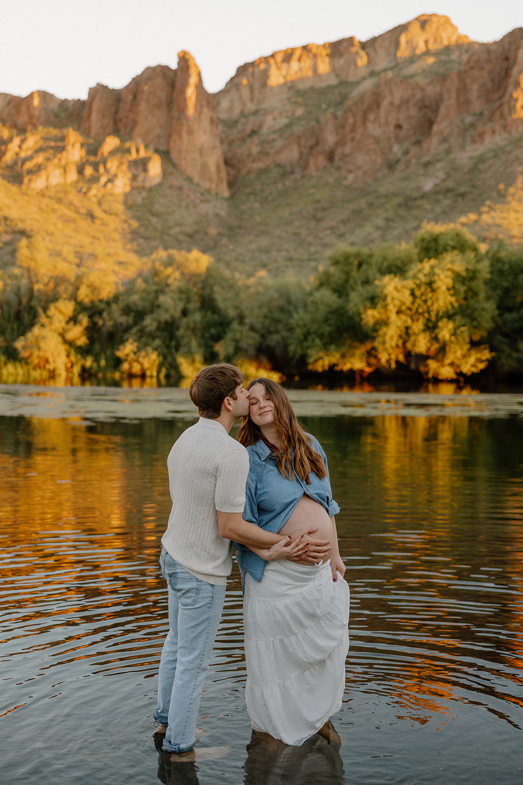 Couple standing in the Salt River at sunset, holding each other with mountains behind them, Arizona maternity photographer
