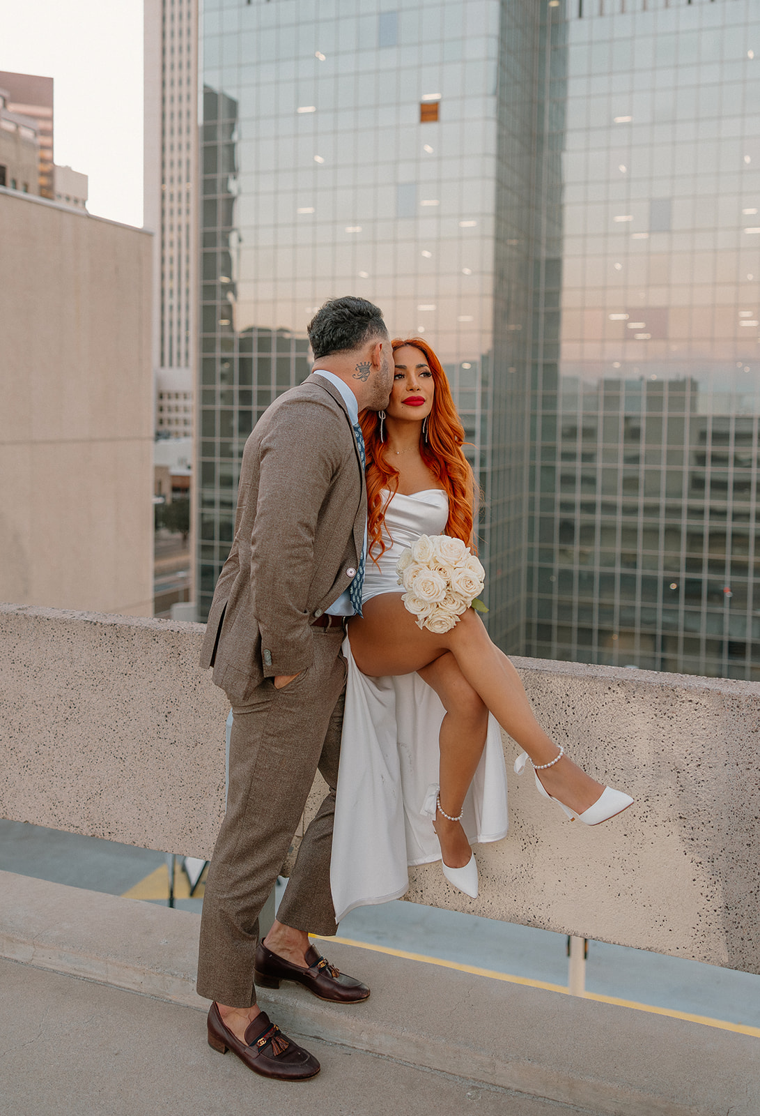 Couple posing on rooftop ledge with downtown skyline during phoenix engagement photos
