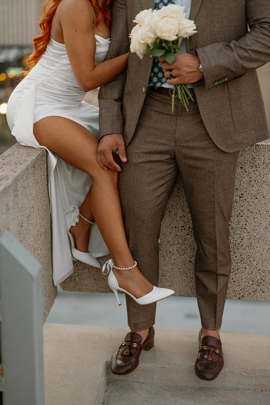 Close-up of couple sitting on rooftop ledge holding white rose bouquet
