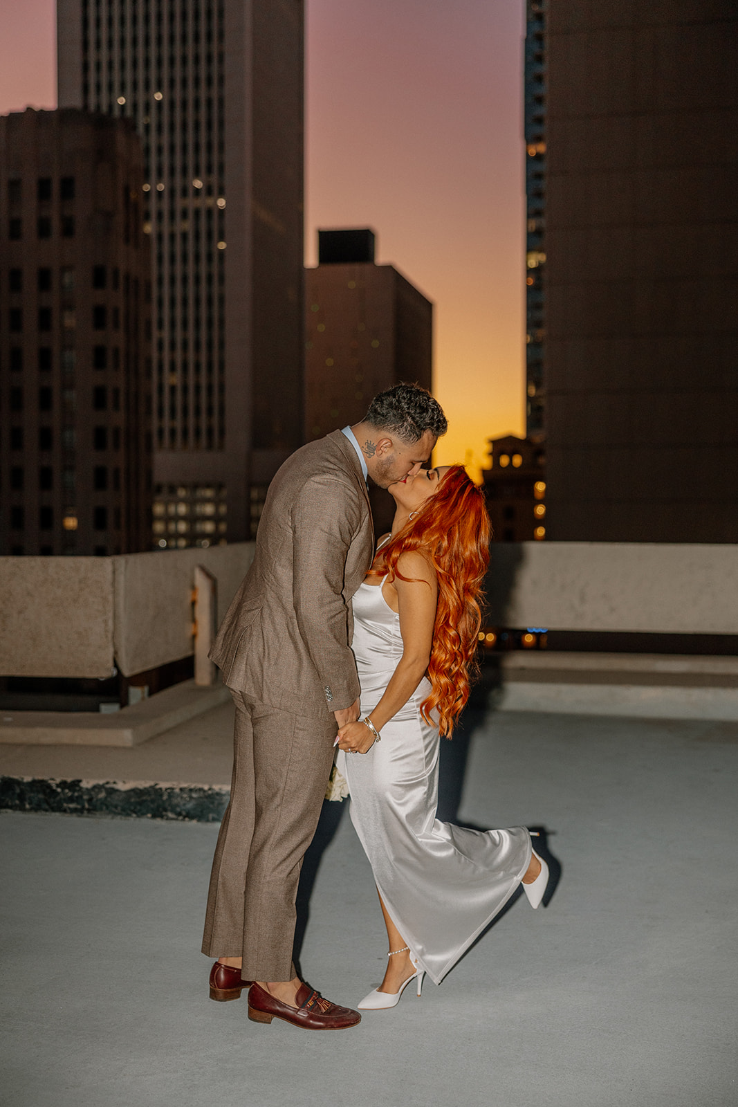Couple kissing on rooftop at sunset with glowing Phoenix skyline behind them
