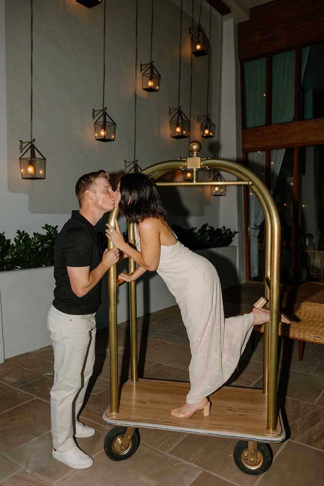 Playful hotel engagement photos of couple kissing on a luggage cart under warm lighting
