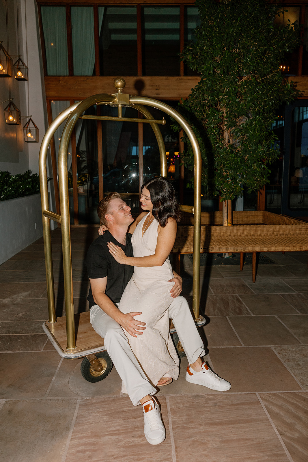 Hotel engagement photos of couple sitting on a luggage cart indoors at night
