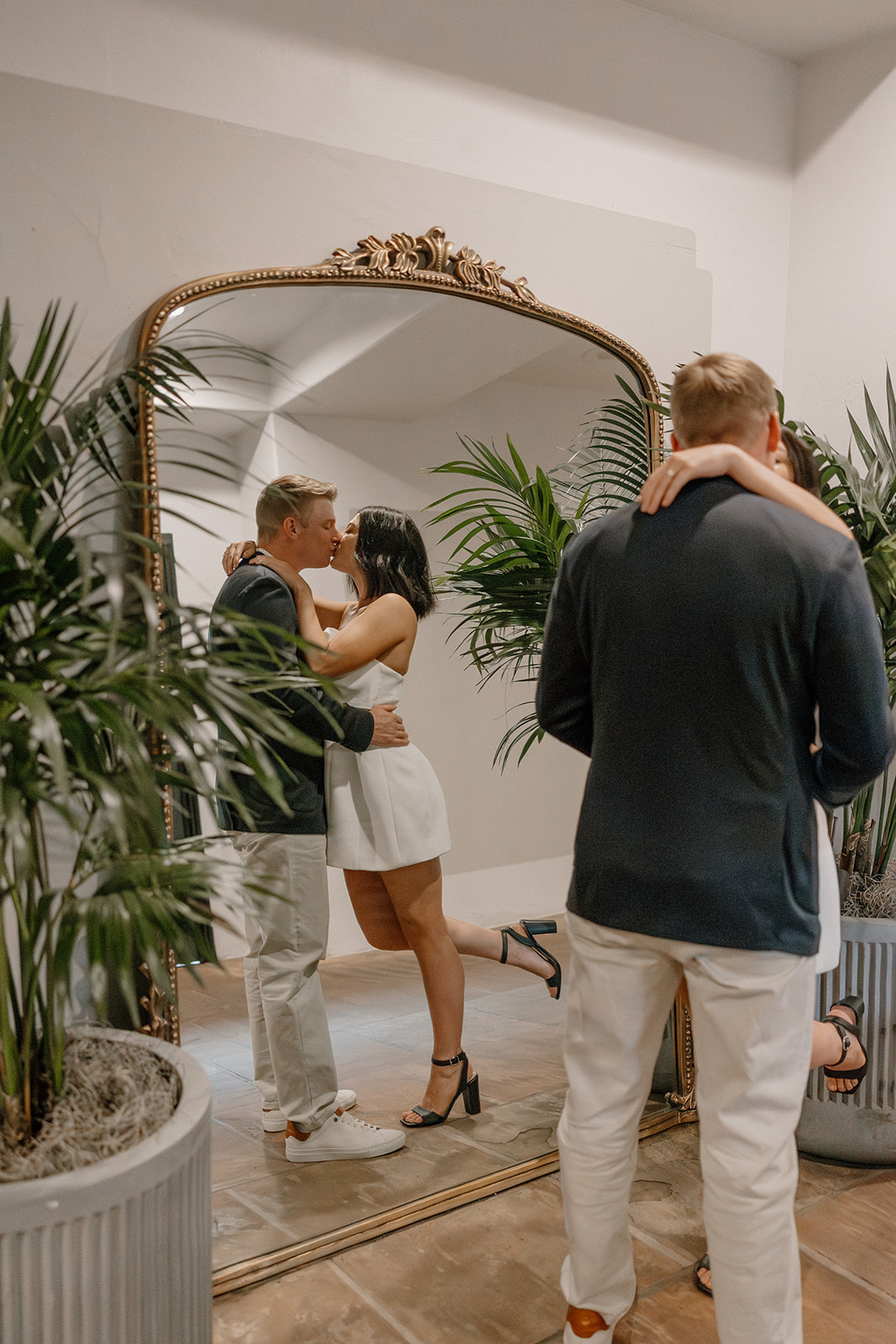 Couple kissing in front of a mirror surrounded by plants during hotel engagement photos
