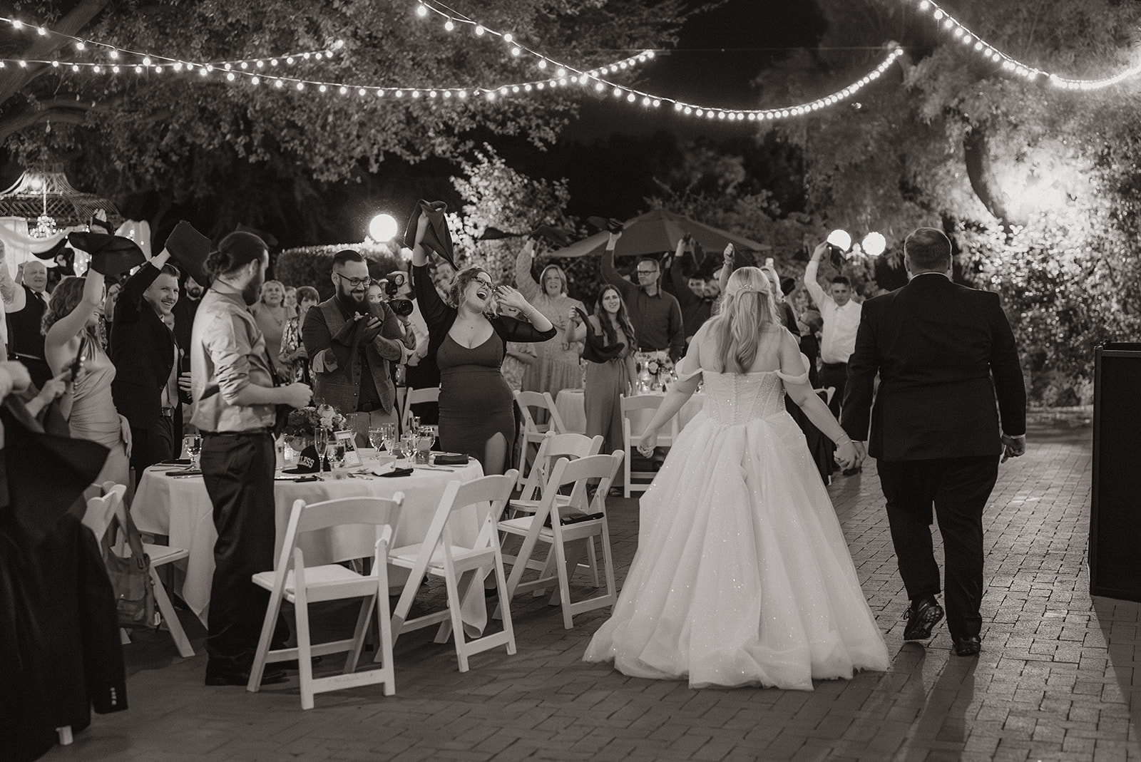 Guests cheering as newlyweds enter their outdoor reception under string lights