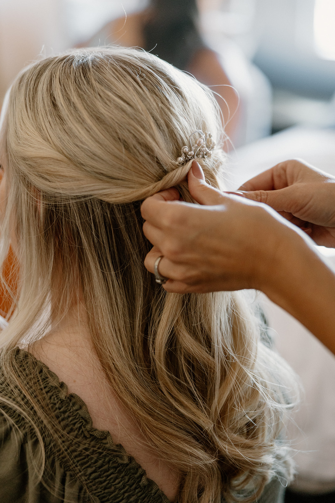 Hairstylist placing pearl hairpiece into bride’s curls during wedding morning
