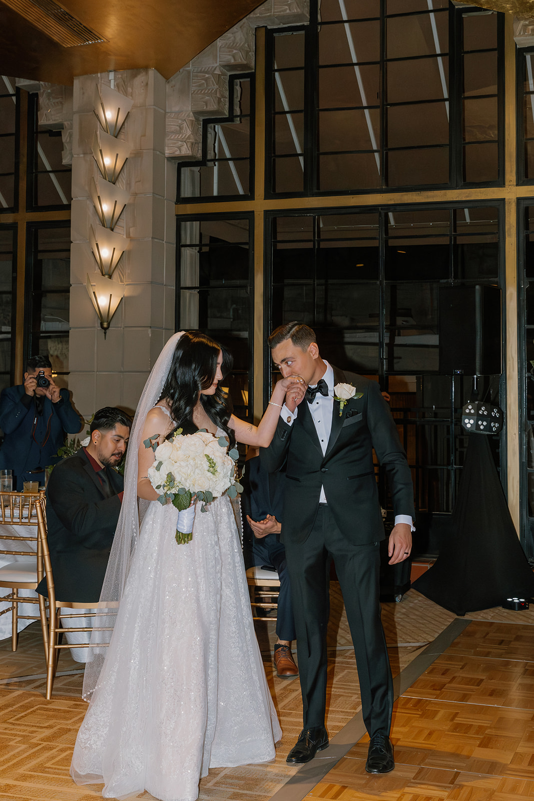 Groom kissing the bride’s hand as they enter the reception space, guests watching from their seats
