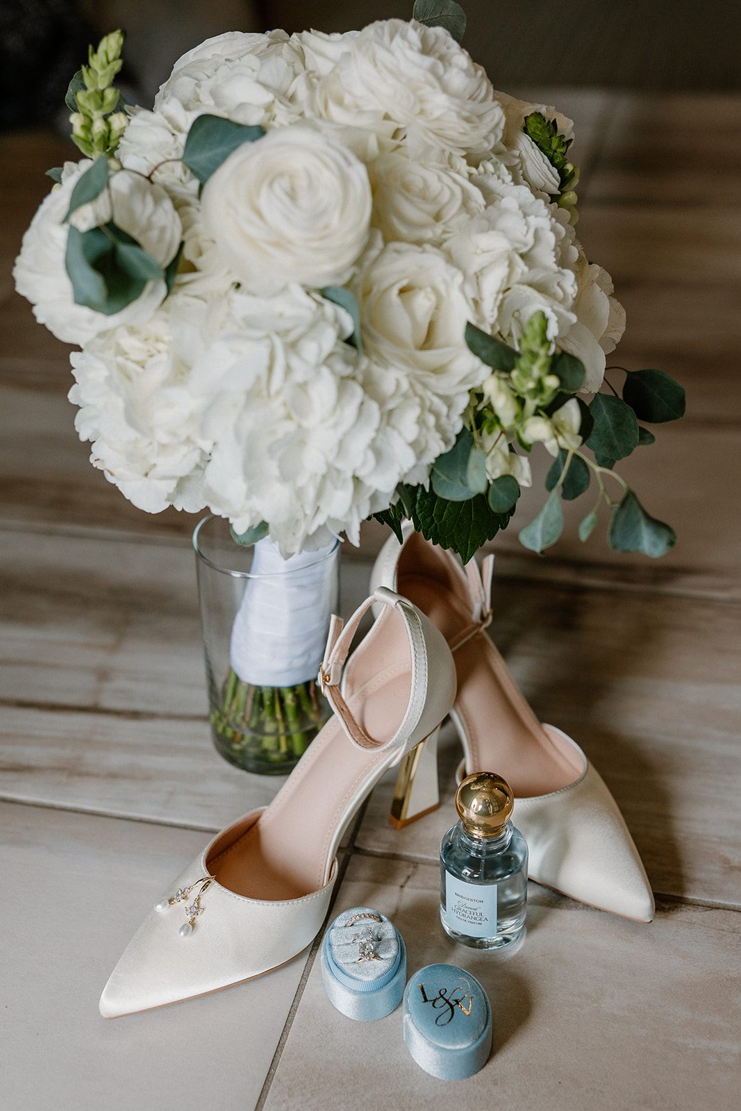 Bridal details with bouquet, shoes, perfume, and ring boxes arranged on a table
