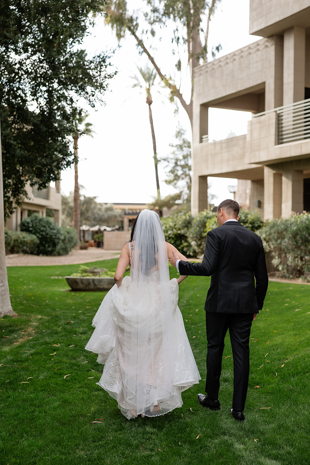 Bride and groom walking away together across the grass with palm trees and resort buildings in the background
