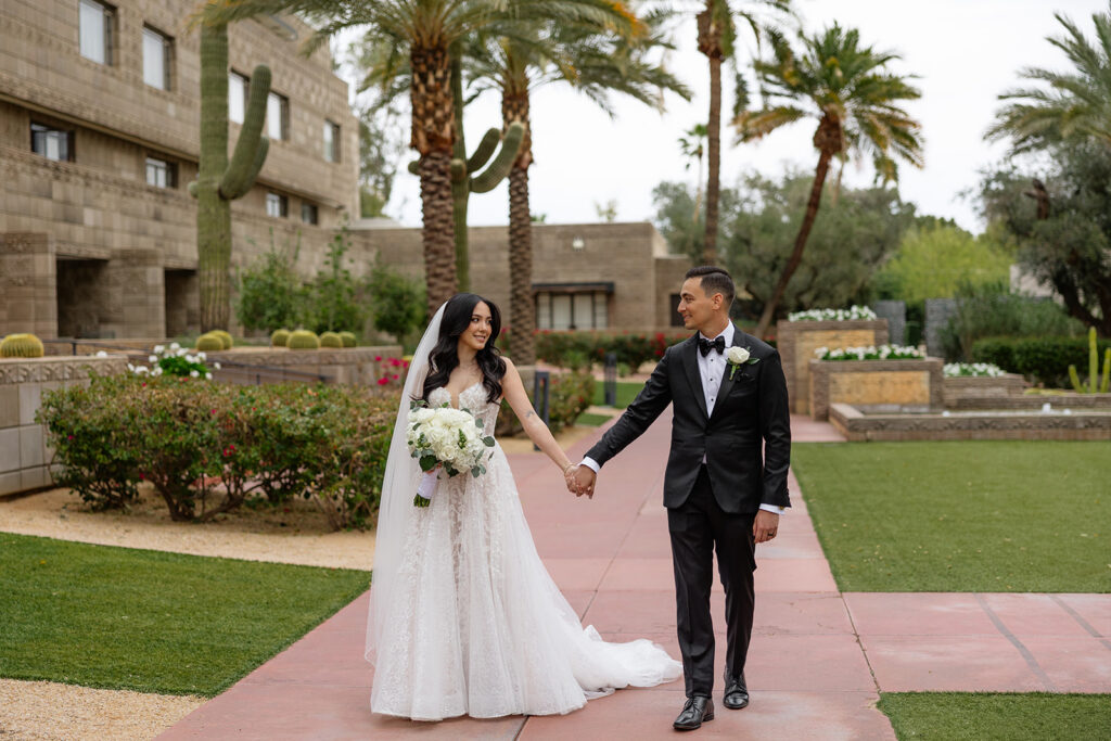 Bride and groom holding hands and walking along a palm-lined path during their Biltmore wedding portraits
