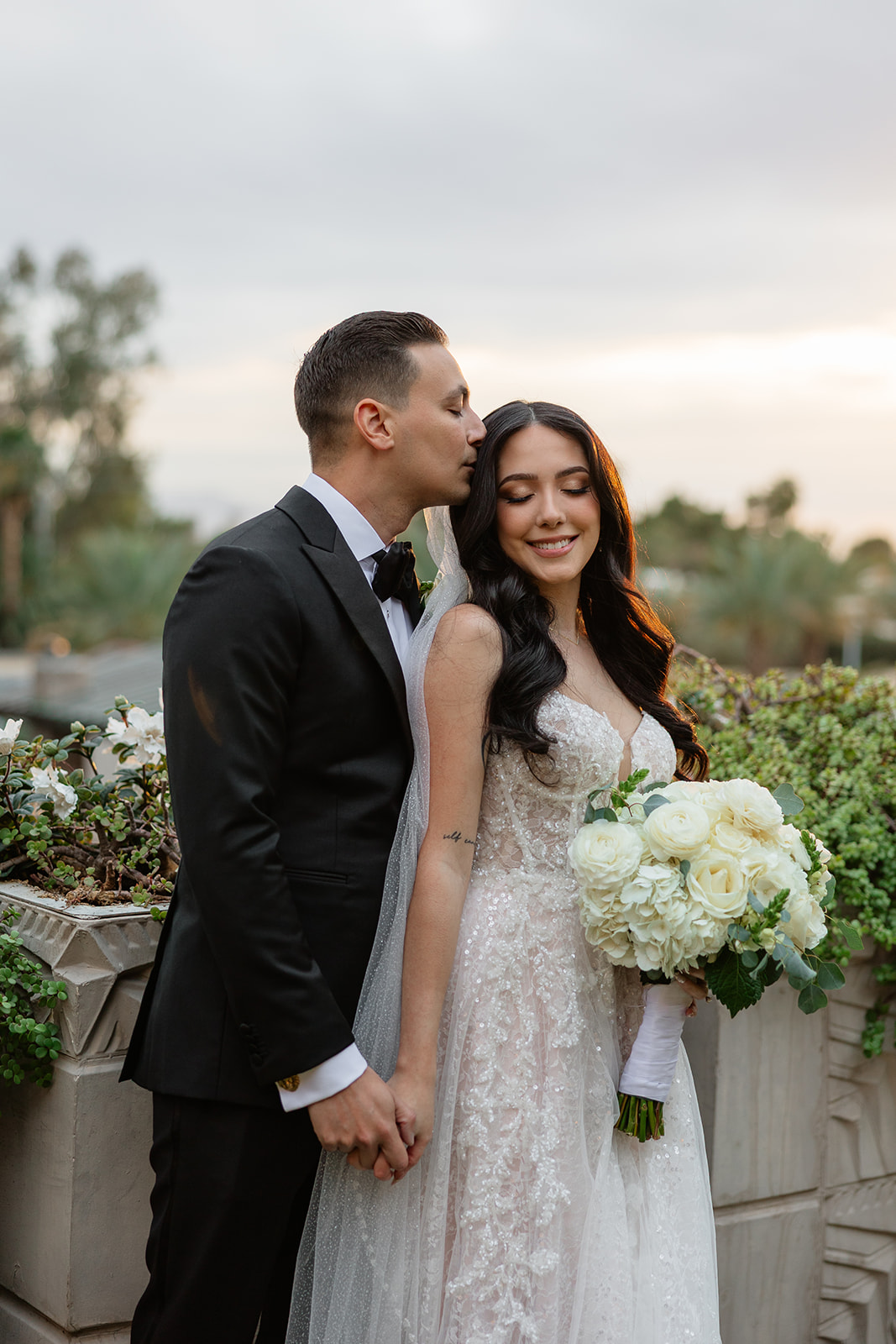 Groom kissing the bride’s temple while they hold hands during golden hour portraits

