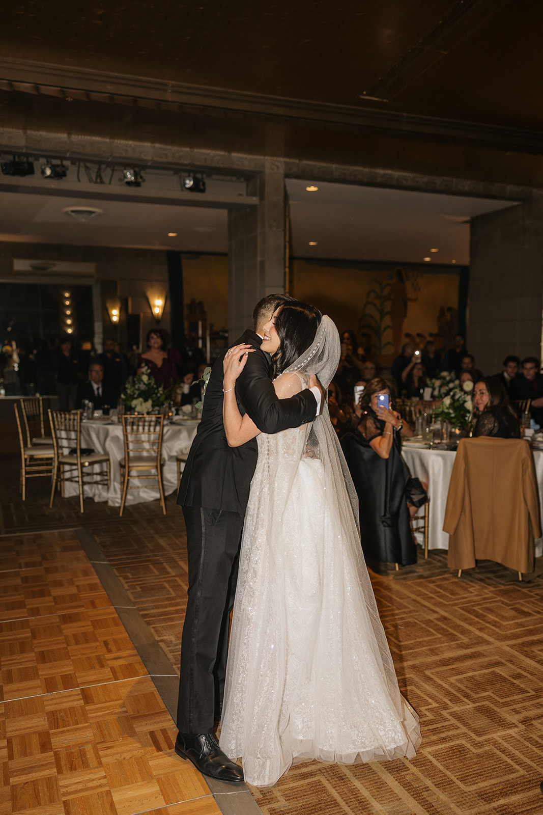 Couple sharing a quiet hug in the middle of the reception as guests watch from their tables
