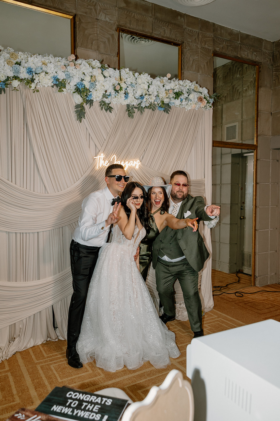 Bride and groom posing with friends in front of a draped floral backdrop and photo booth setup
