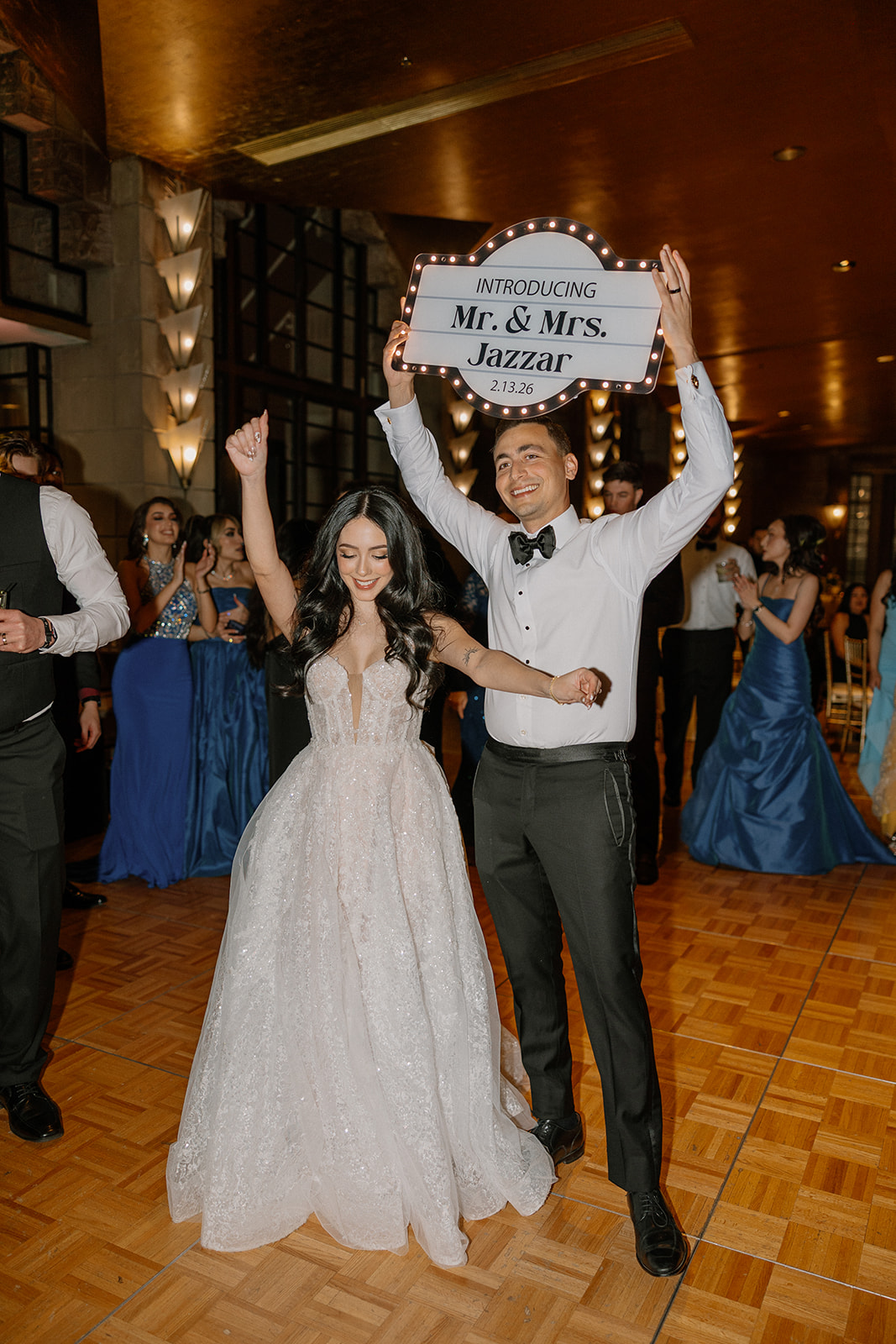 Bride and groom entering the reception with a sign reading “Introducing Mr. & Mrs.” while guests cheer
