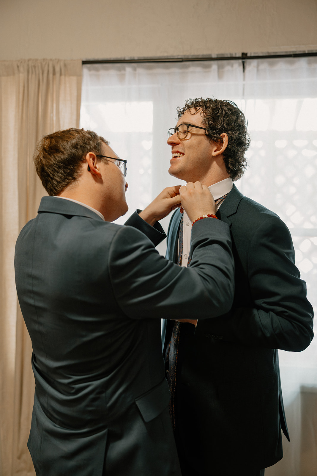 Groomsmen helping groom get ready in suit near window light before Arizona wedding
