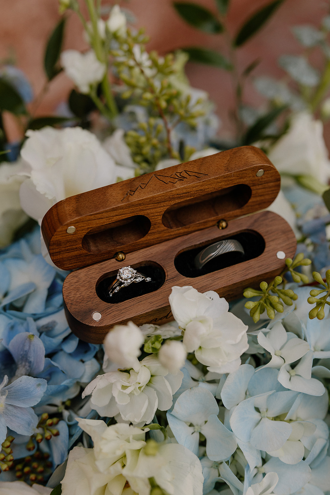 Wedding rings in wooden box resting on blue and white floral bouquet detail shot
