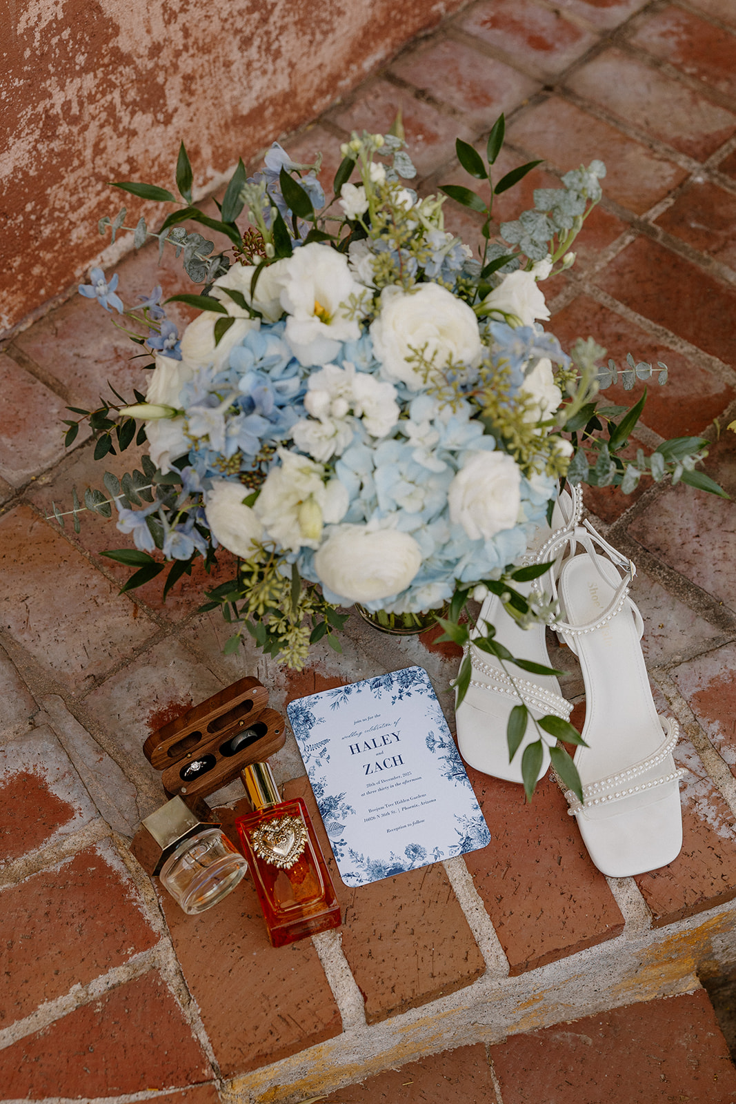 Bridal bouquet with blue and white flowers, wedding invitation, shoes, and perfume on terracotta tile floor
