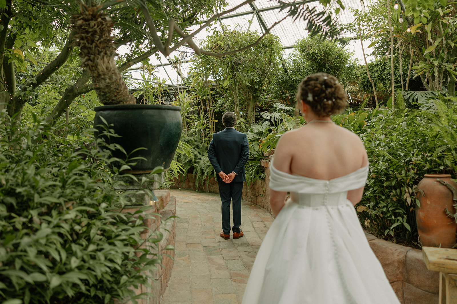 Bride walking toward groom for first look inside lush greenhouse wedding venue with tropical greenery
