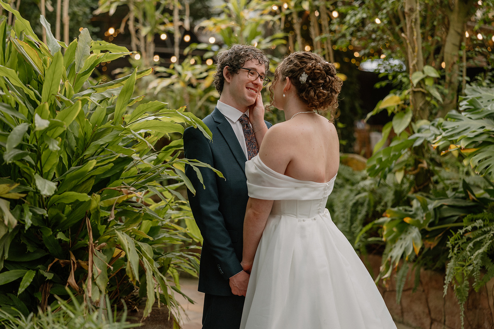 Couple sharing an intimate moment surrounded by greenery and string lights inside a romantic greenhouse setting
