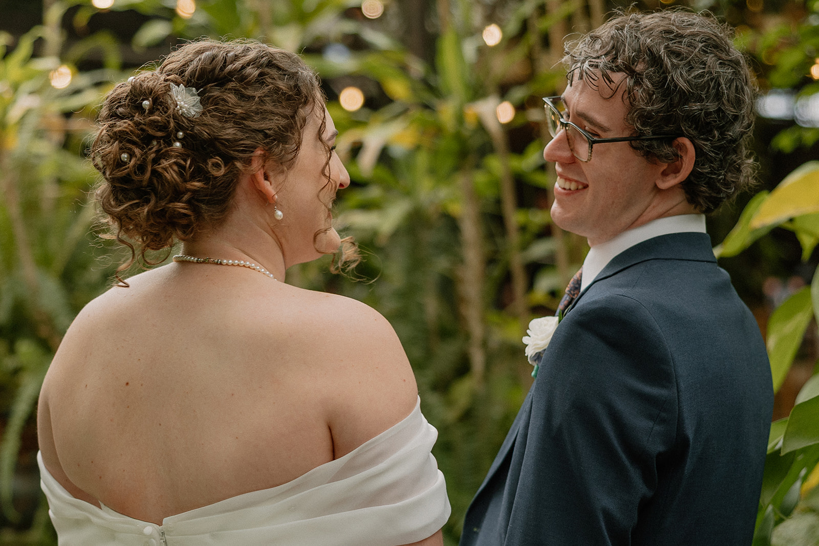 Newlyweds smiling at each other in a tropical garden setting with string lights and greenery
