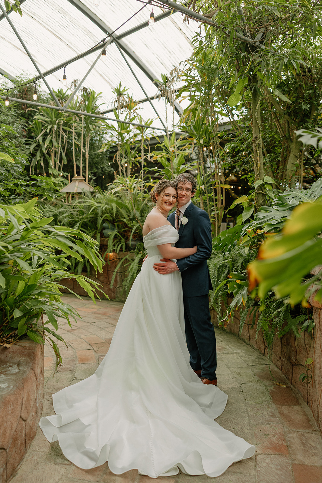 Bride and groom posing together in greenhouse-style Arizona wedding venue filled with tropical plants
