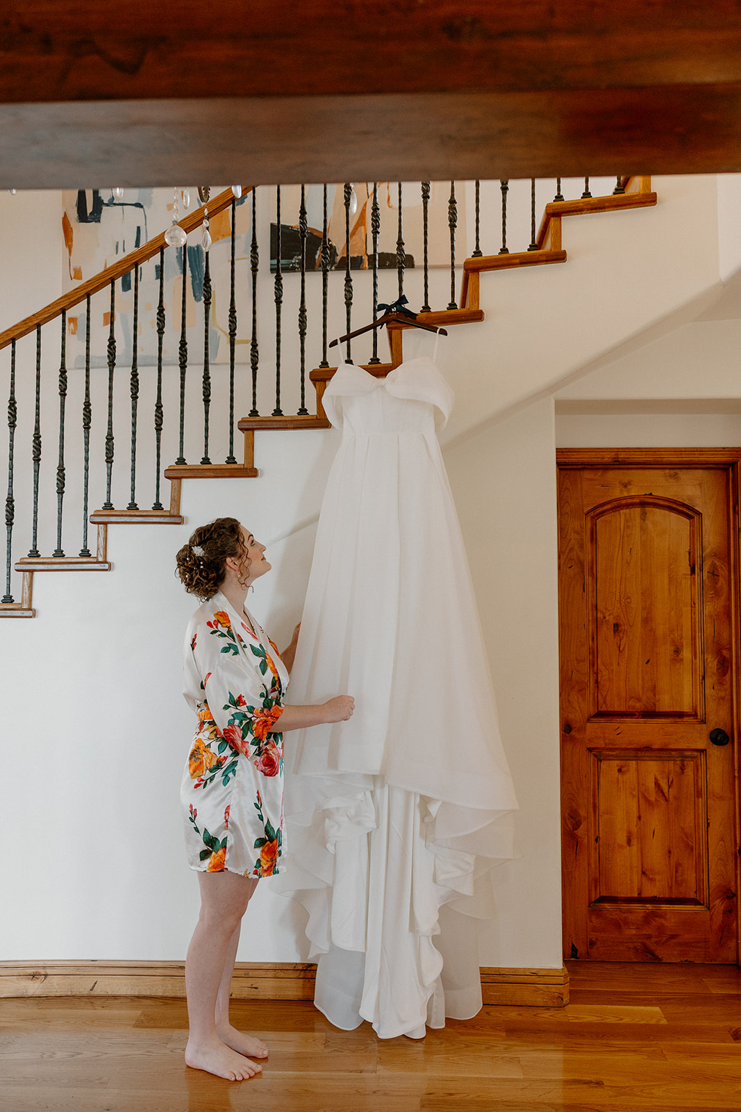 Bride admiring off-the-shoulder wedding dress hanging on staircase inside Arizona home
