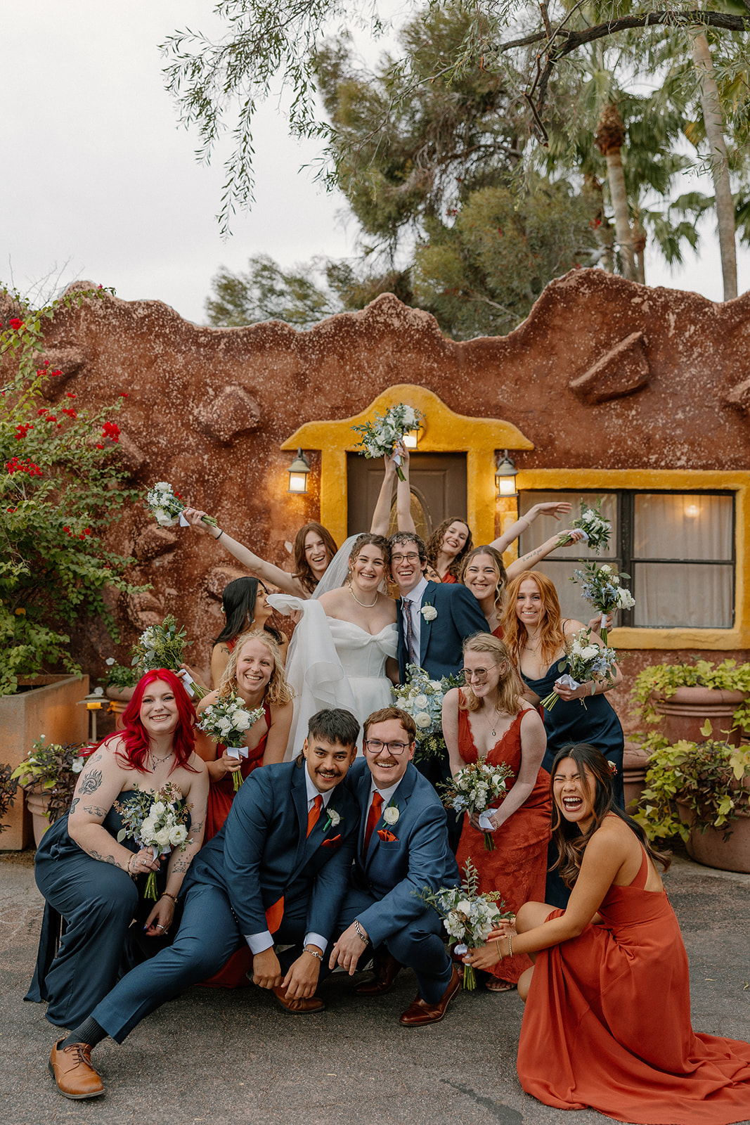 Wedding party group photo in front of rustic adobe building at Arizona wedding venues with colorful florals
