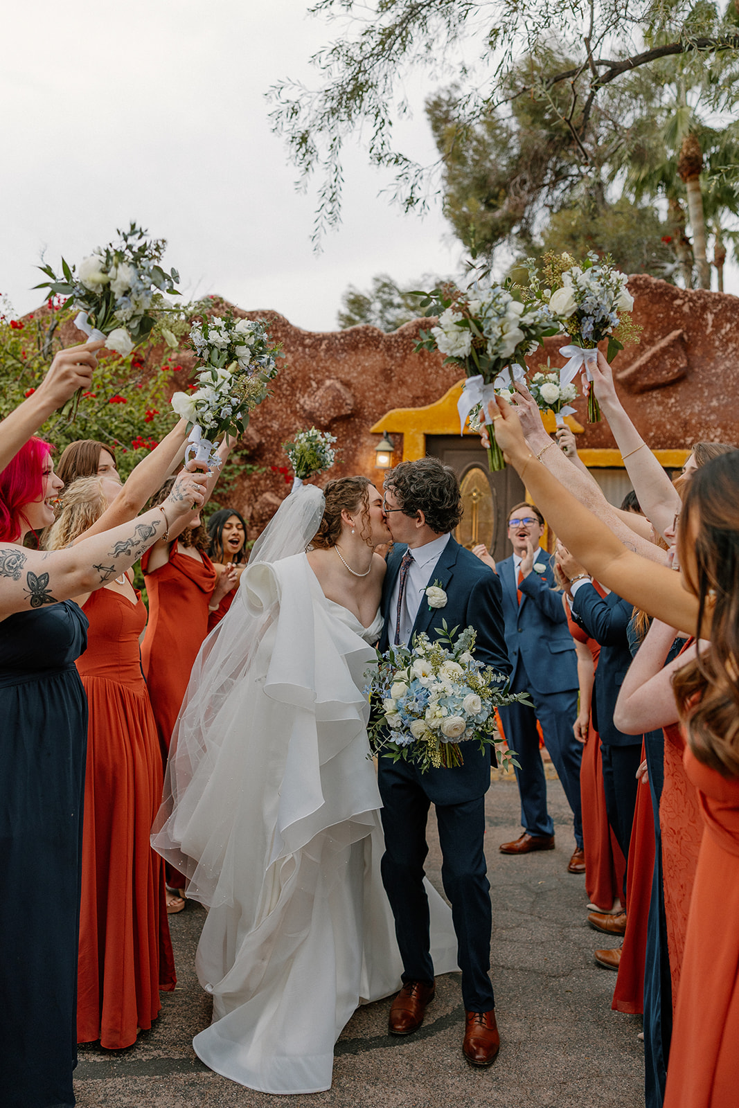 Newlyweds kissing under raised bouquets outside adobe-style Arizona wedding venue with bridal party cheering
