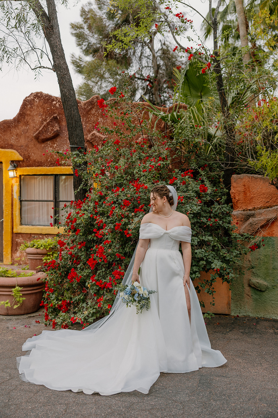 Bride standing with bouquet in front of vibrant red flowers and adobe-style architecture at an Arizona wedding venue
