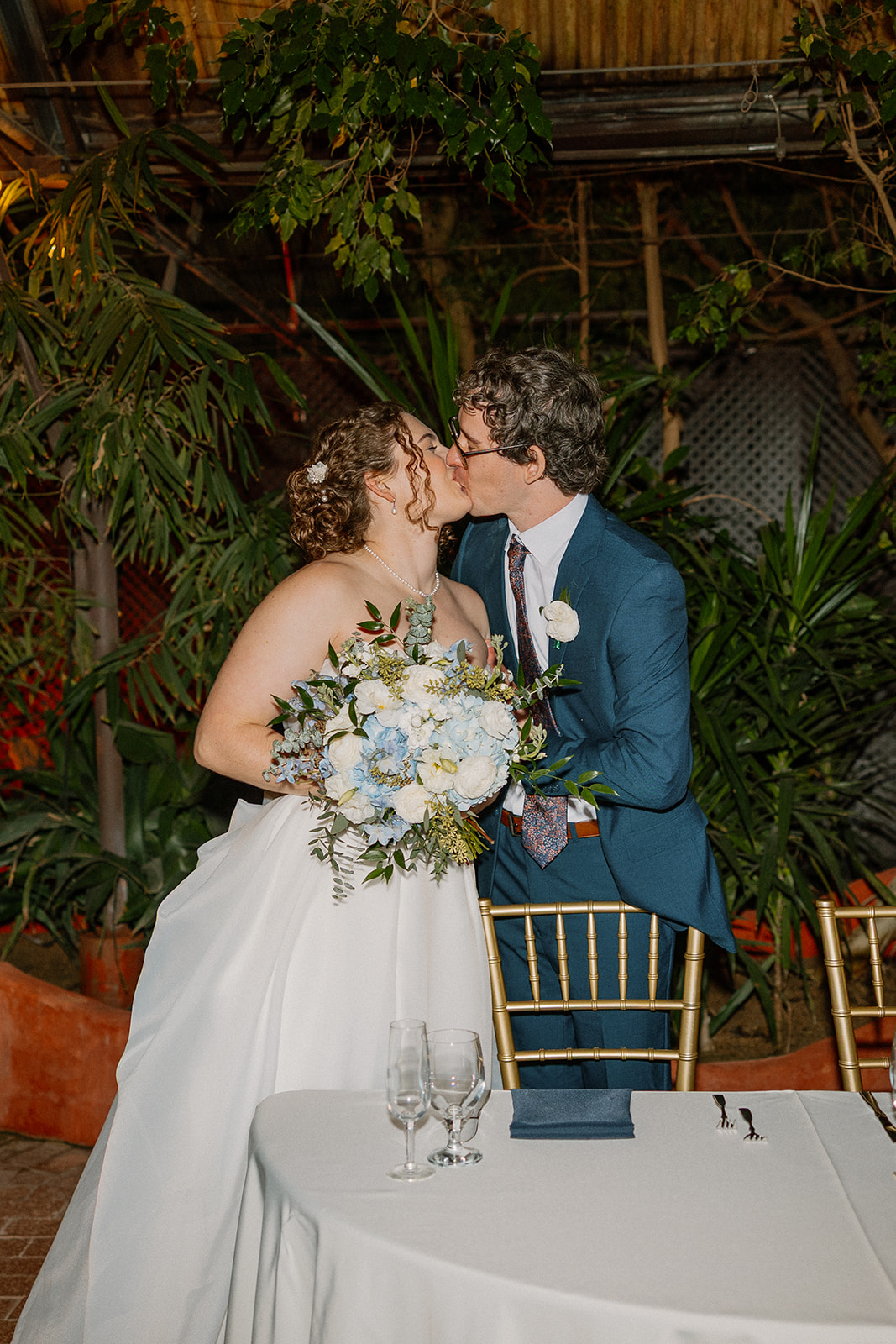 Bride and groom kissing at sweetheart table surrounded by greenery and soft reception lighting
