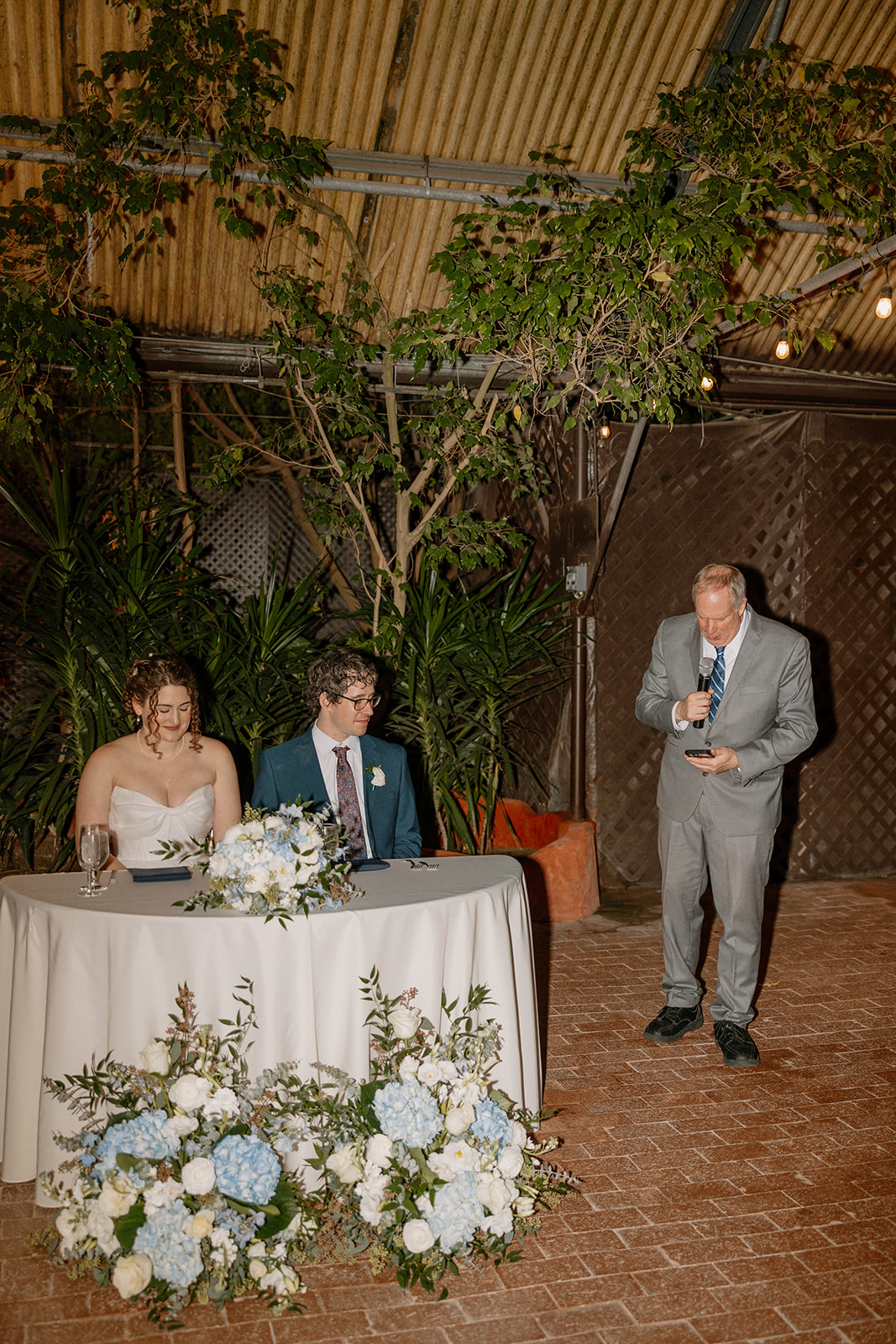 Bride and groom seated at sweetheart table during reception speech at an Arizona wedding venue with lush greenery backdrop
