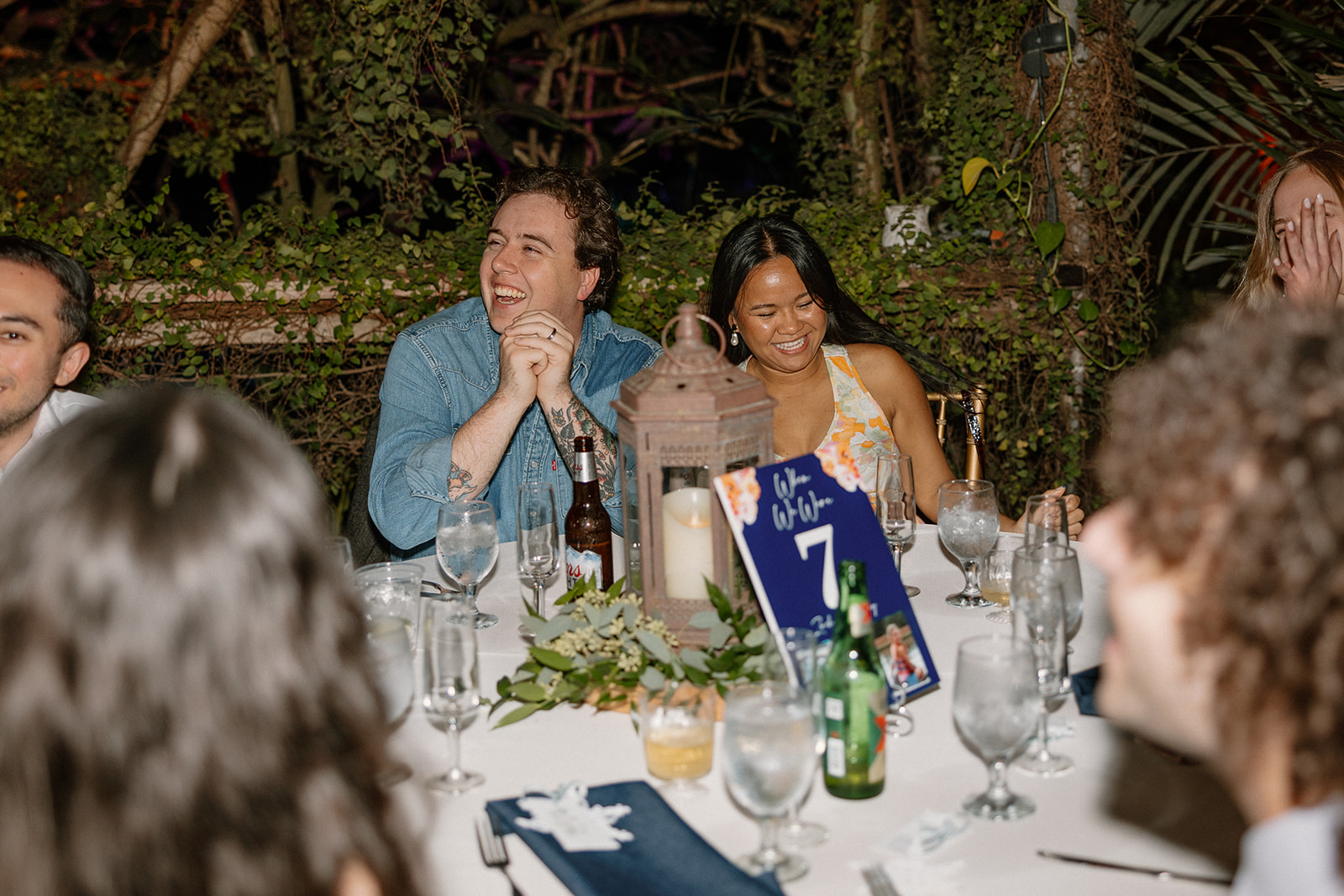 Wedding guests laughing at reception table with candles, greenery, and drinks in a cozy indoor setting

