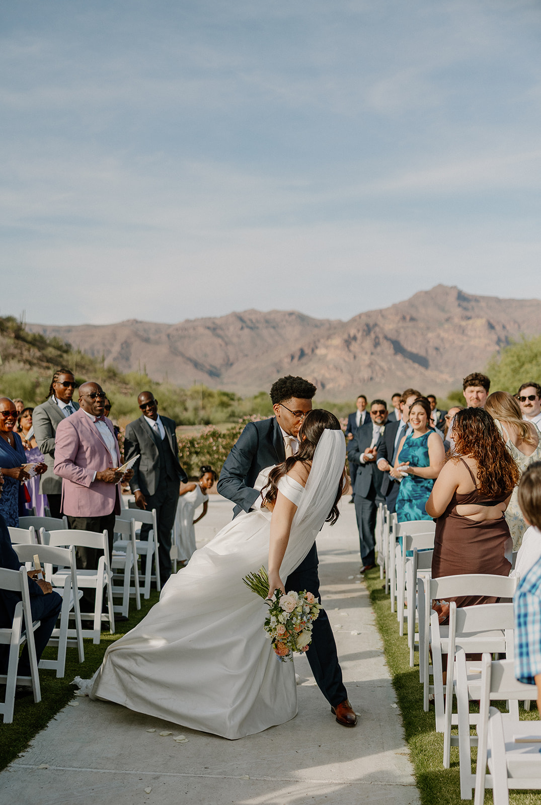 bride and groom kissing in aisle after ceremony with guests watching and mountains behind them at gold canyon golf resort wedding